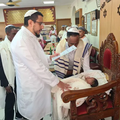 המוהל איתמר חמו בחלוק לבן קורא ברכה ליד כיסא אליהו והתינוק, הסנדק עטוף בטלית