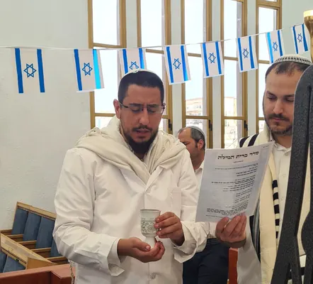 המוהל איתמר חמו בחלוק לבן ובטלית מברך על כוס היין בברית, דגלי ישראל ברקע