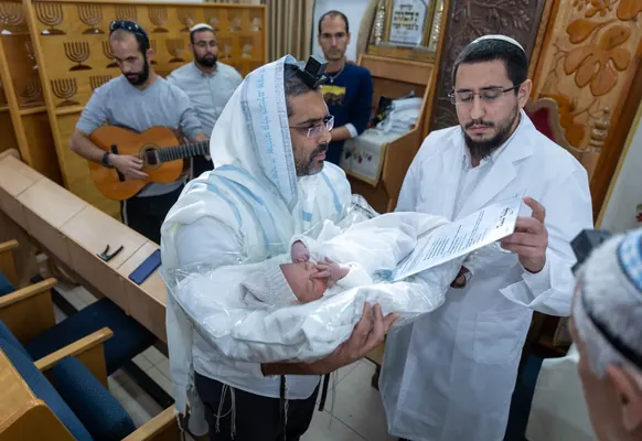 איתמר חמו קורא את הברכה ליד הסנדק שמחזיק את התינוק, נגן גיטרה מאחור